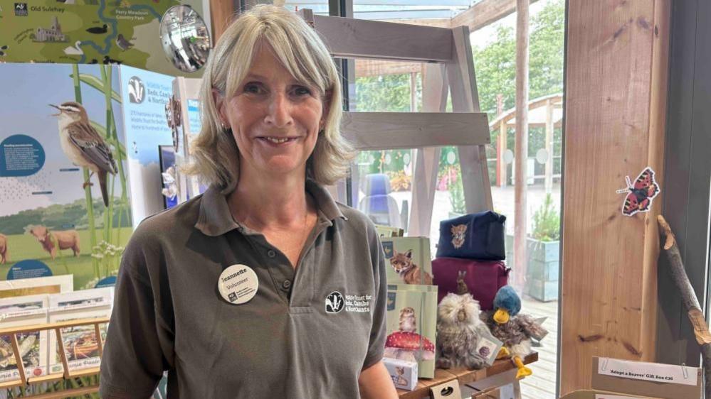 Jeanette Smith, wearing a brown polo shirt, standing in a shop, smiling and looking at the camera. She has shoulder length fair hair. She is buy items to  purchase. 