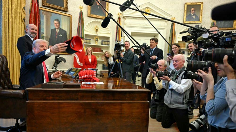 Image shows Trump in the Oval Office with pool reporters in front of desk