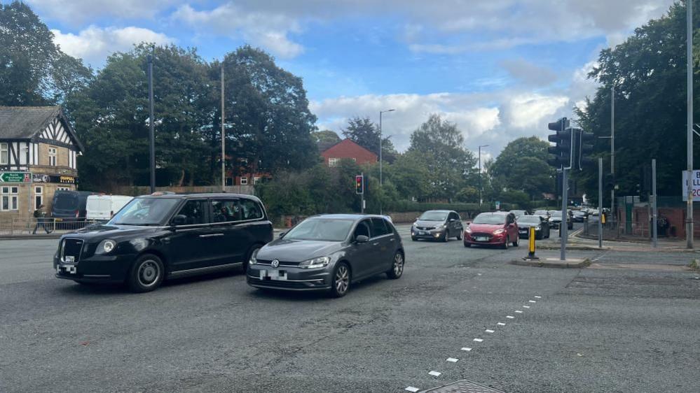 Two lanes of traffic drive along on a busy road at a junction with another road. 