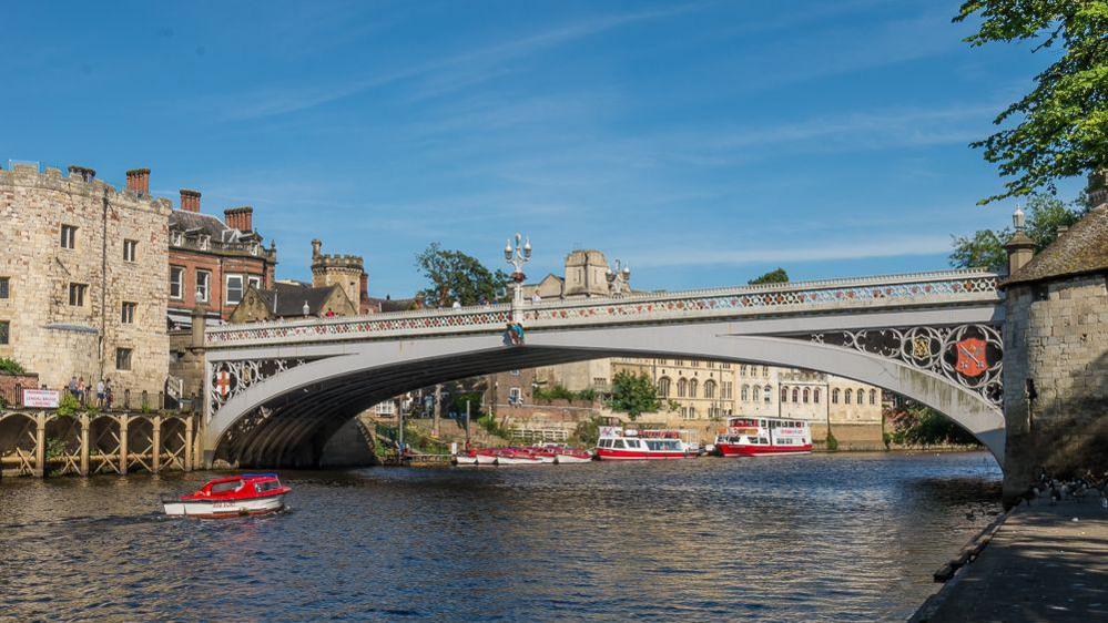 Lendal Bridge, an iron bridge with Gothic influences, over the River Ouse in York. A handful of small red boats can be seen bobbing in the water, alongside two bigger vessels.