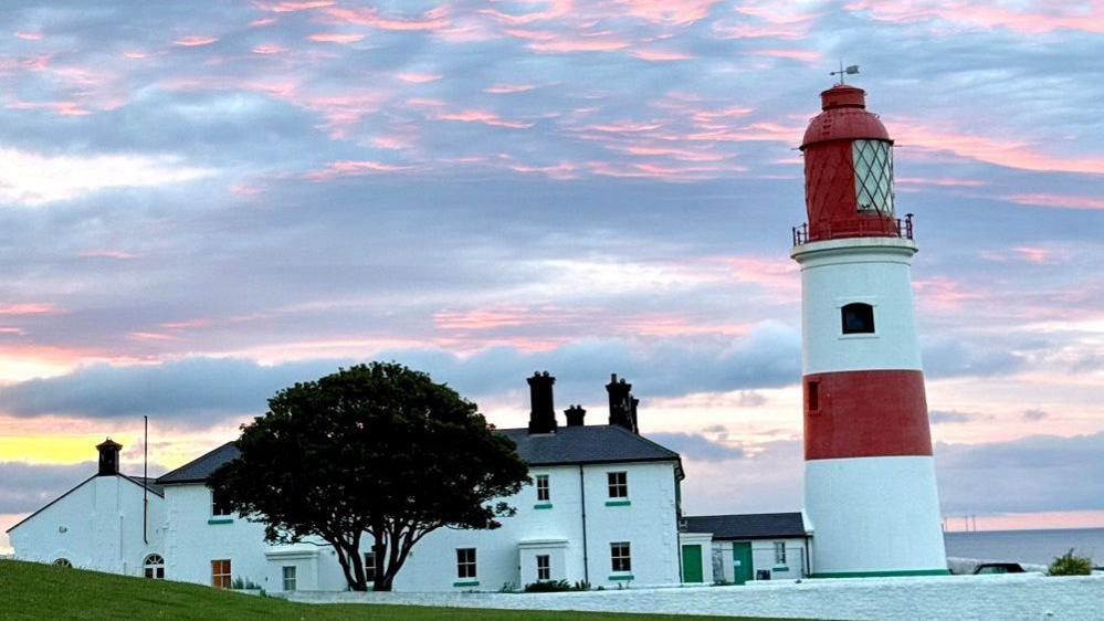 Souter lighthouse which is large with red and white stripes. Besides it is a white, stone building. The sky is turning grey with bits of pink in it.