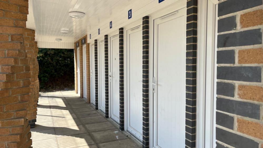 A row of doors, with numbers above on a brick building, with buff bricks and black pillars and corbelling. The pillars on the veranda frontage have ofset twist patterns. Paving flags are on the ground.