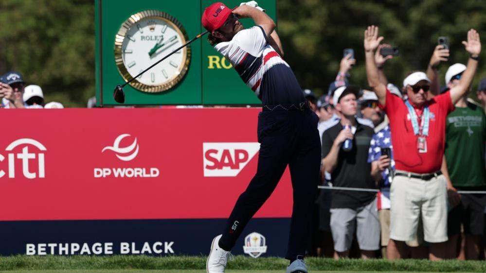 Cameron Young of team USA on the 6th hole during the singles matches of the 2025 Ryder Cup golf tournament. 

He is in the final stage of swinging his golf club. He is wearing the Team USA kit - white shoes, navy trousers, a golf t-shirt which is made up of blue, red and white stripes and a red cap with the words USA on the front. In the background the crowd watches to see where the ball lands.