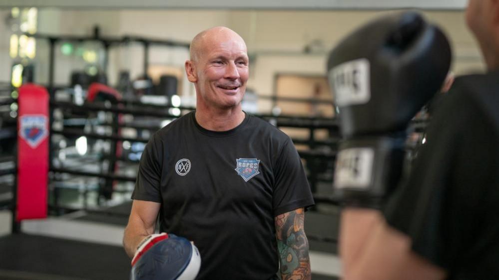 A bald man wearing a black top with tattoos on his left arm. He wears one boxing pad on his right hand and smiles at a blurred individual in the foreground. A boxing ring can be seen in the background.