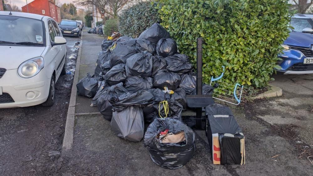 Black bin bags piled up on the pavement next to a white car and green hedge. 