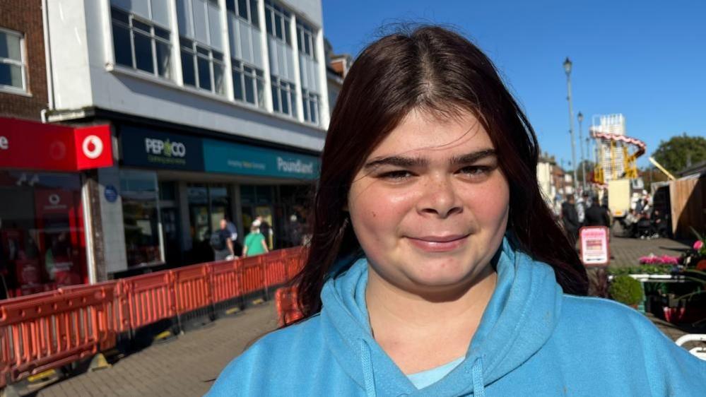 Amy Newey smiles directly at the camera. She has long black hair and is wearing a blue hoodie. She is standing near Poundland in Great Yarmouth where there are orange pedestrian barriers in the street owing to groundworks.