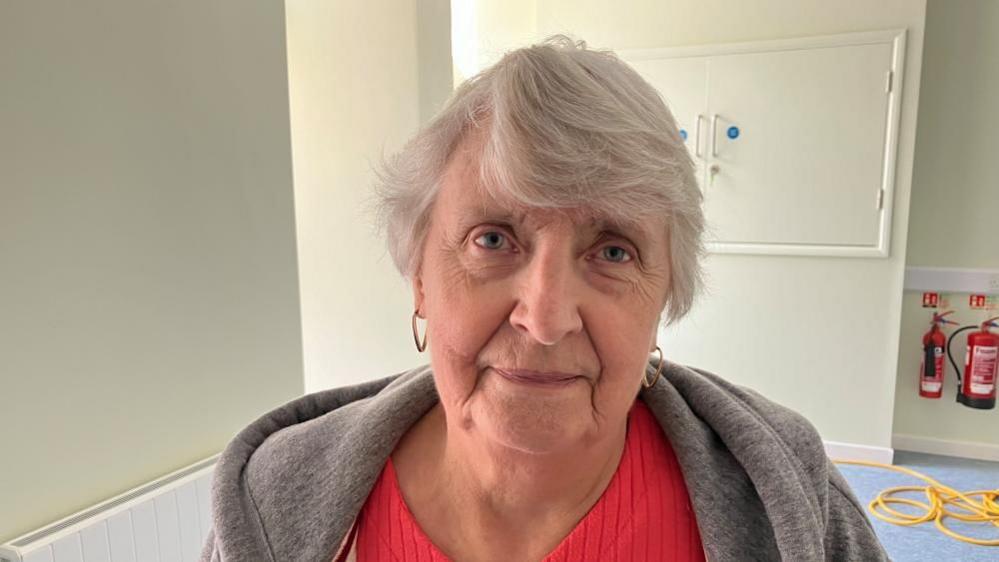 Carol Wilson with medium-length grey hair and large gold earrings. She is wearing a red top and grey coat. There is a white cupboard and fire extinguishers behind her.