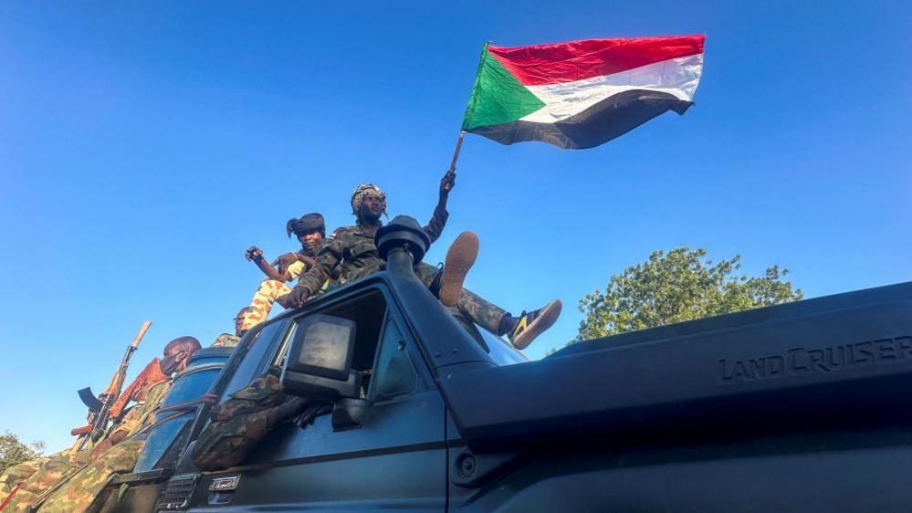 Men in camouflage,  one of whom is waving a flag, are in a land cruiser in Wad Madani in Sudan on 12 January 2025