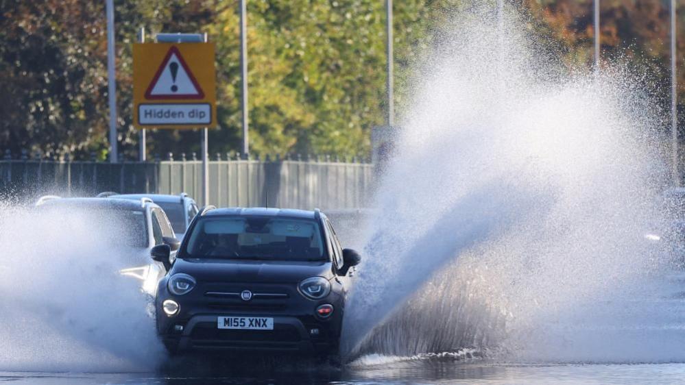 A car driving through flood water causing a large amount of spray either side of the car