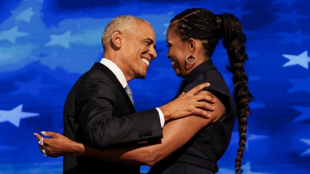 Barack and Michelle Obama facing each other and hugging. Barack has short grey hair and is wearing a black suit. Michelle has long black hair and is also wearing a black dress. They are standing in front of a blue background with stars.