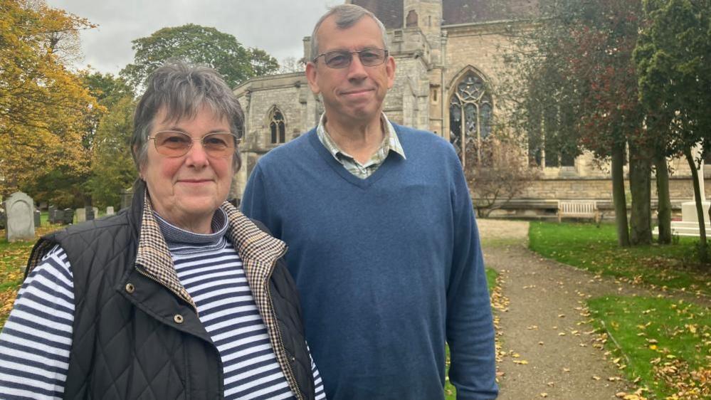 Caroline and Roger Wandless stand outside St Helen Church in Escrick. Caroline has short grey hair, and wears glasses, a blue and white striped top and a black gilet. Roger has a blue jumper and a green checked shirt.