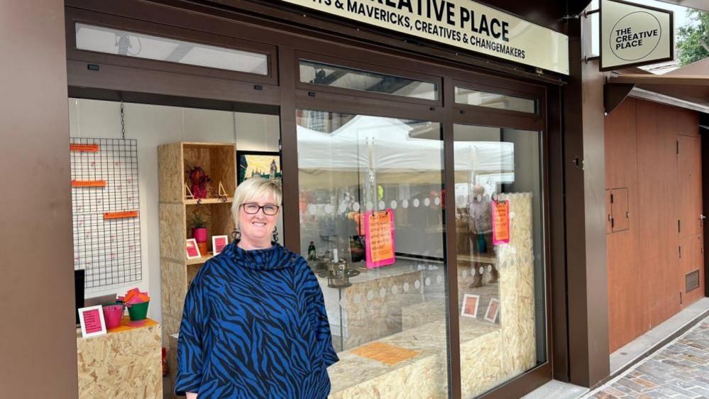 Becky Carrier with short straight blond hair and brown glasses wearing a blue and black dress stands outside a brown market stall with display shelves and a "creative place" sign.