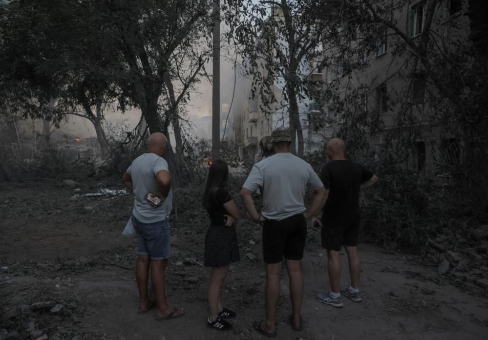 Three men in shorts and tee shirts and a young woman in a summer dress stand in a yard and look at smoking rubble after an attack in Kramatorsk blamed on the Russian military.
