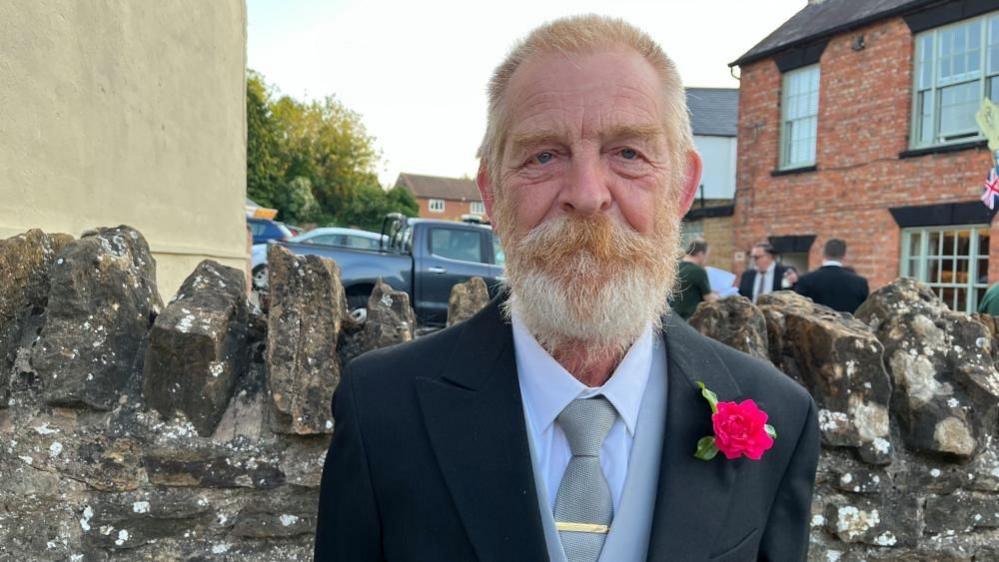 Frank in a dark-coloured suit, tie and waistcoat, standing in front of a brick wall. He is wearing a flower pinned to his jacket lapel. He has a thick beard.