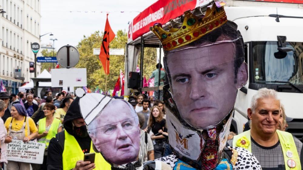 At the front of a crowd of protesters, two hold up cardboard cut-outs of Macron wearing a crown and Barnier wearing a papal hat