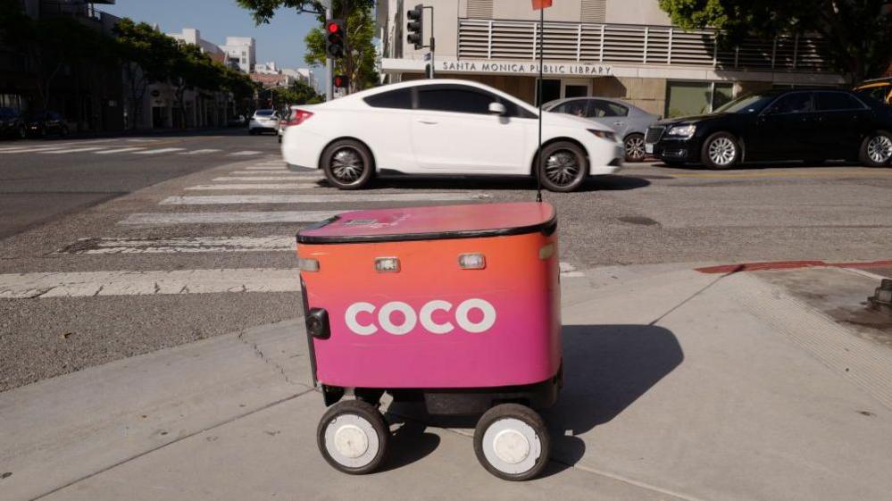 A delivery robot in front of a car on a street in Los Angeles