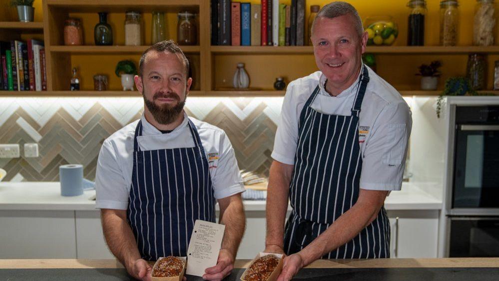 Two chefs in a show kitchen wearing striped aprons and each holding a small cake