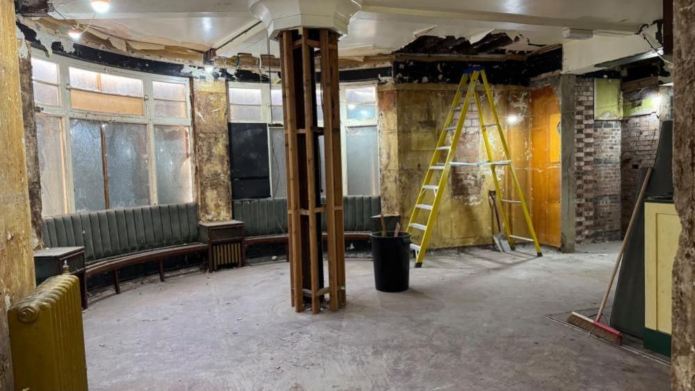 A curved seat partially removed, and a column clad with timbers to support a finished surface. Brickwork has been exposed, and a ladder stands in the corner. 