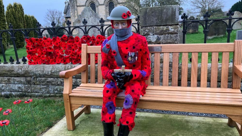 A statue of a man covered in red and purple poppies wearing a helmet, wrapped up in a scarf, with war medals pinned to his chest, sitting on a wooden bench outside a church.