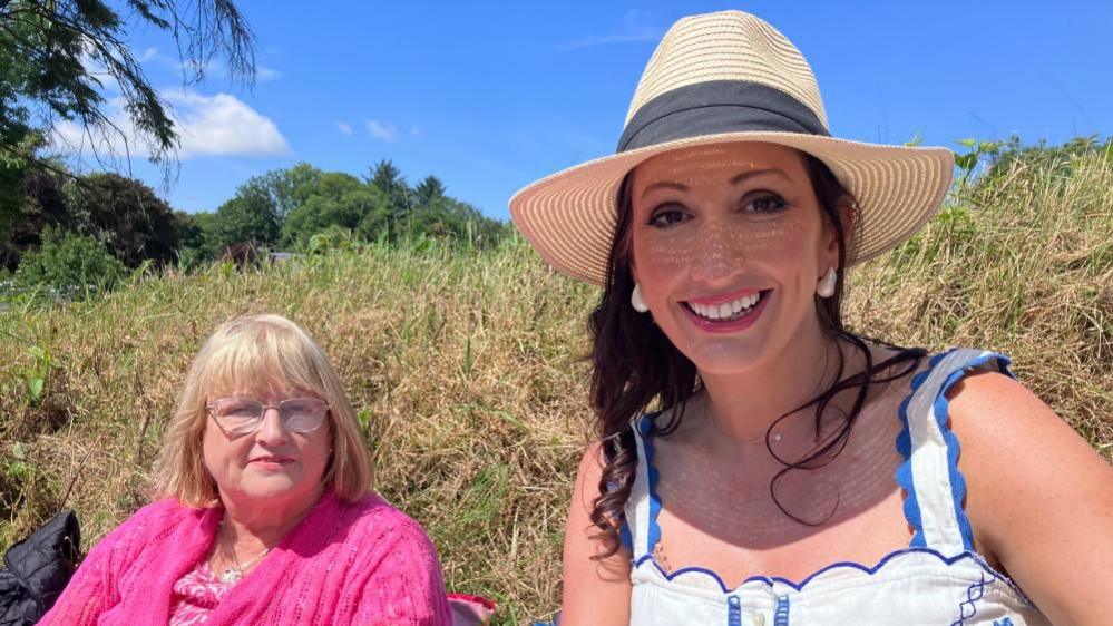 Two women smile at the camera. On the left, Maureen Little has short blonde hair and is wearing a pink top. On the right Emma Little-Pengelly is smiling and wearing a straw hat and a white and blue vest top. she has long brown hair.