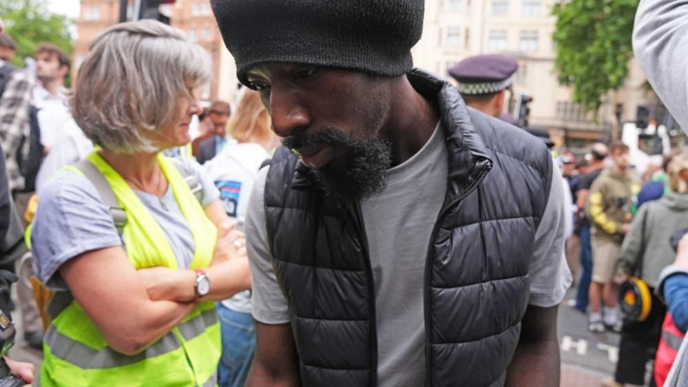 Charles, with a goatee and wearing a grey t-shirt, a black quilted gillet, and a black beanie walks through a large crowd into court.