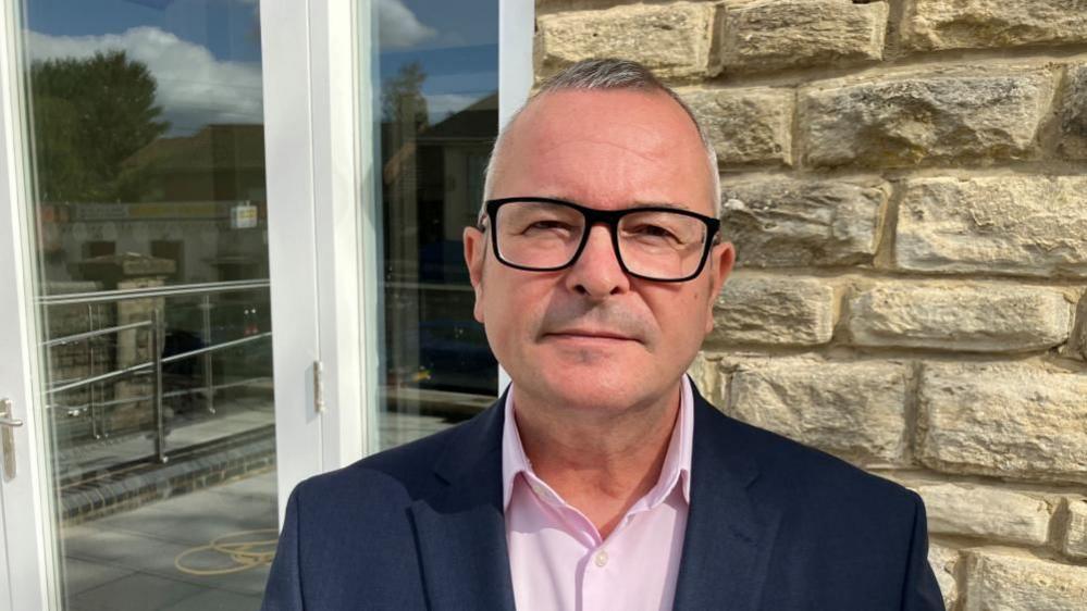 Lee Barron with short white hair and black-rimmed glasses wearing a dark blue jacket and pink shirt, standing in front of a stone wall and PVC glass door.