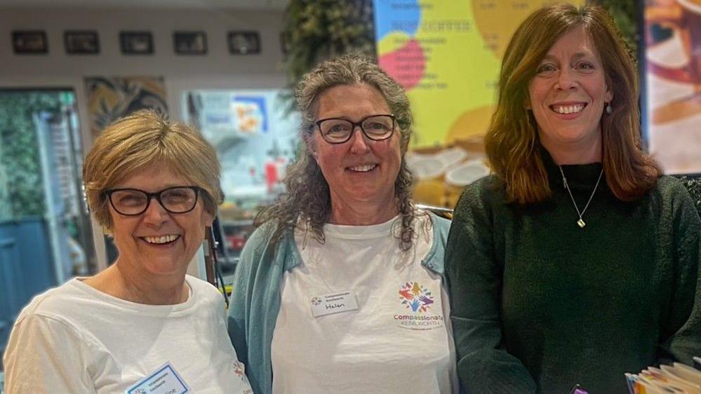 Three women stood inside a community hub with bright posters and leaflets smiling at the camera 