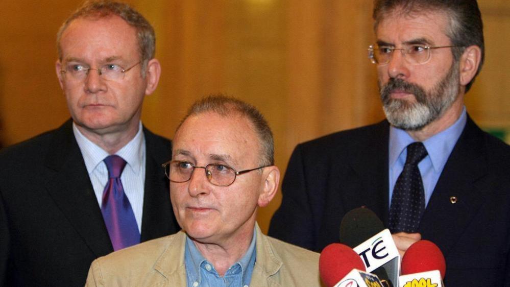 Martin McGuinness, Denis Donaldson and Gerry Adams are all looking to their right. All three are wearing glasses. Mr McGuinness and Mr Adams are both wearing dark suits, shirts and ties. Mr Donaldson is wearing a tanned coloured jacket and a denim shirt. There are three microphones in front of them.