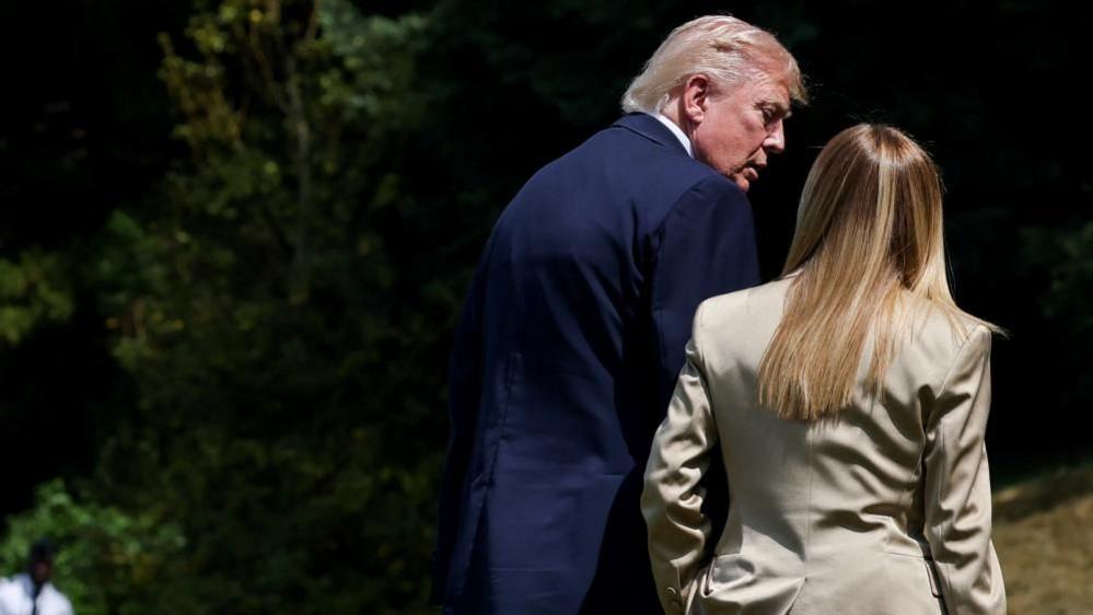 A man in a blue suit talks with his back to the camera to a woman with blonde hair and a beige suite, again with her back to the camera