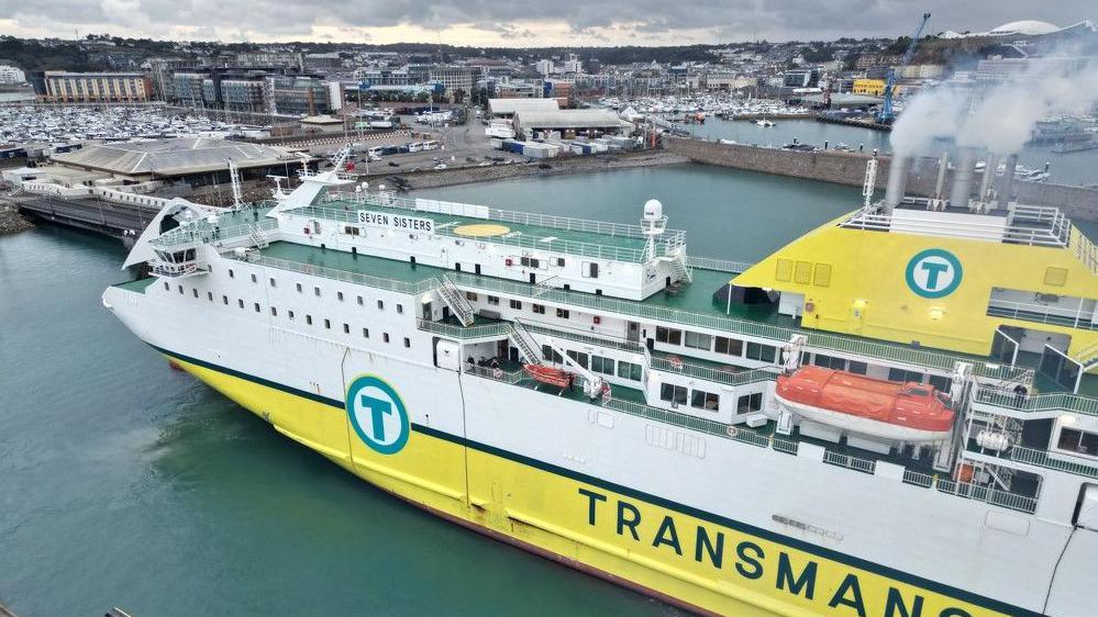 A ferry enters St Helier Harbour. Its lower half is yellow. The word 'Transmanche' is painted in green  letters on the side. The upper half is white. An orange lifeboat lies on an upper deck. Smoke billows from chimneys atop the vessel. 