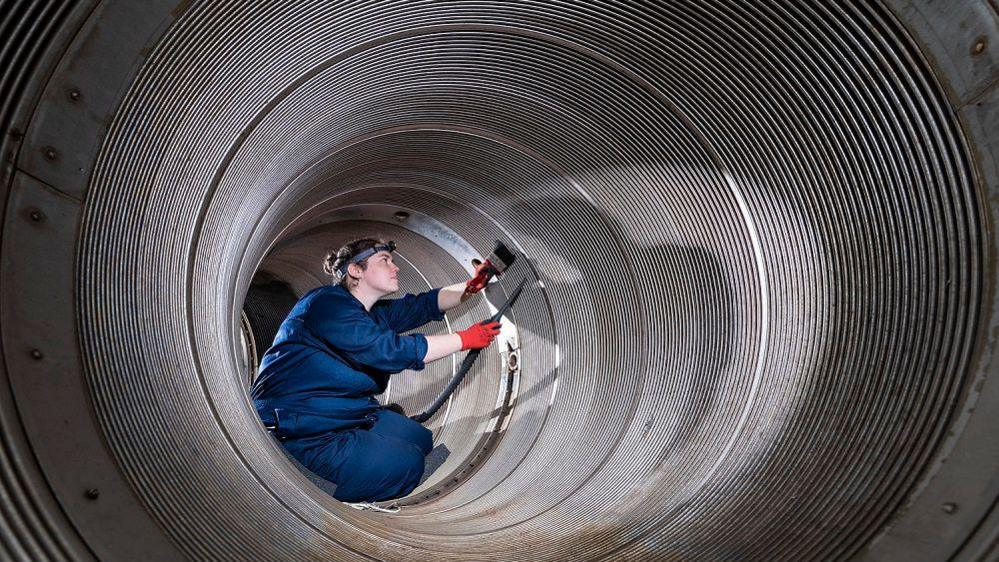 A woman with headtorch, gloves, brush and specialist vacuum wearing a blue boiler suit sits inside a large metal tube, cleaning it.