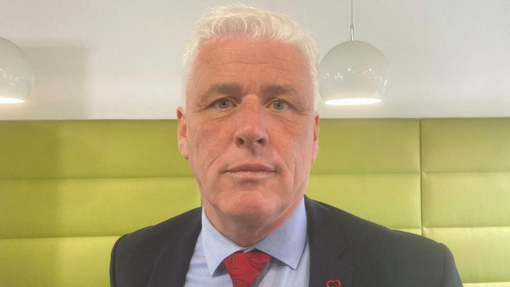 Fearghal McKinney has short white hair and is wearing a dark grey suit jacket, a light blue shirt and a red tie. He has a heart shaped pin on the right side of the blazer. 