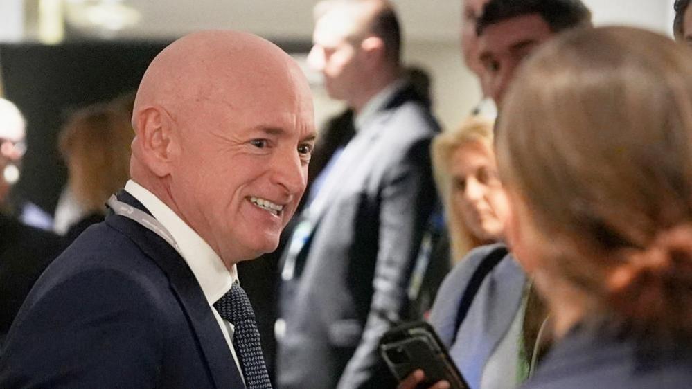 Senatori Mark Kelly, me një kostum blu të errët dhe kravatë me motive, buzëqesh dhe kthehet nga kamera para një grupi njerëzish.