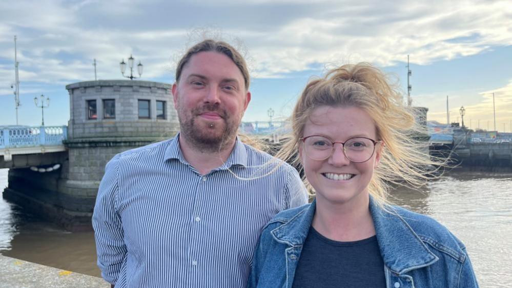 Aaron Bennett, wearing a blue striped shirt. His hair is tied back, he's bearded and has a pierced lower lip. Georgia is standing to the right. She has blonde hair, blowing in the wind, and is wearing glasses, a blue t-shirt and blue denim jacket. They are standing next to the River Yare in Great Yarmouth, with Haven Bridge in the background.