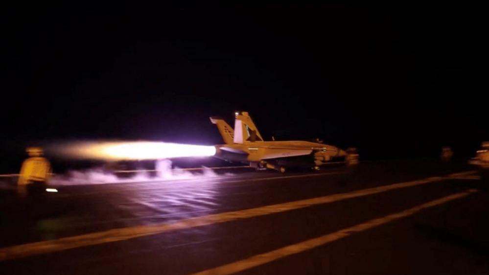 A large flame blasts from the back of a fighter jet on a runway with an air traffic controller stood nearby