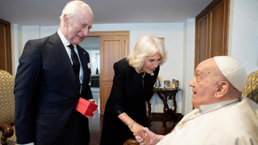 King Charles III pictured standing next to Queen Camilla who is shaking Pope Francis's hand while the Pope is seated