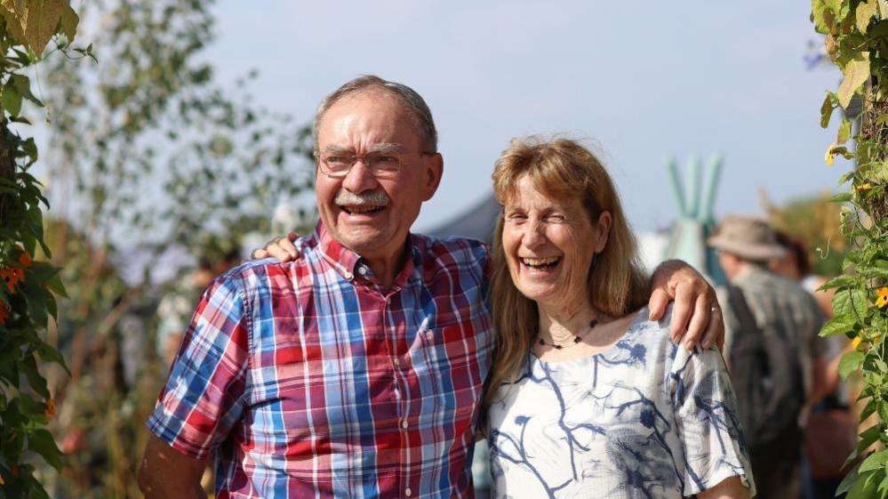 a man wearing a red and blue checked shirt smiles as he has his arm around a woman with shoulder-length hair and a white and blue patterned t shirt. 