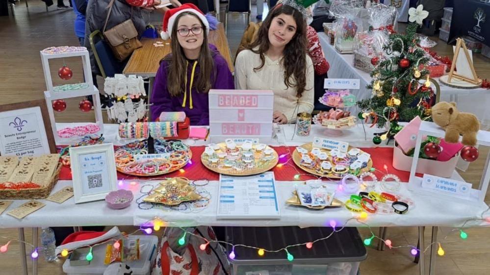 A girl with long brown hair, glasses, a santa hat and a purple hoodie and a girl with long brown hair and a cream jumper - sit behind a table with a white table cloth and plates with bracelets. A light box says Beaded Beauty.