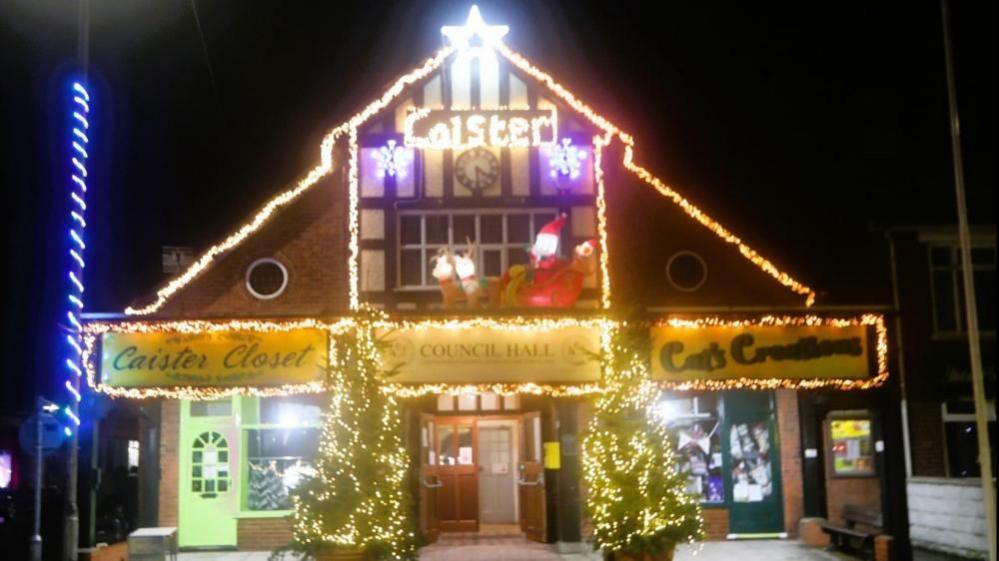 Caister Council Hall decorated with Christmas lights, with two Christmas trees, and a street light decorated with a twisted rope braid of blue lights.