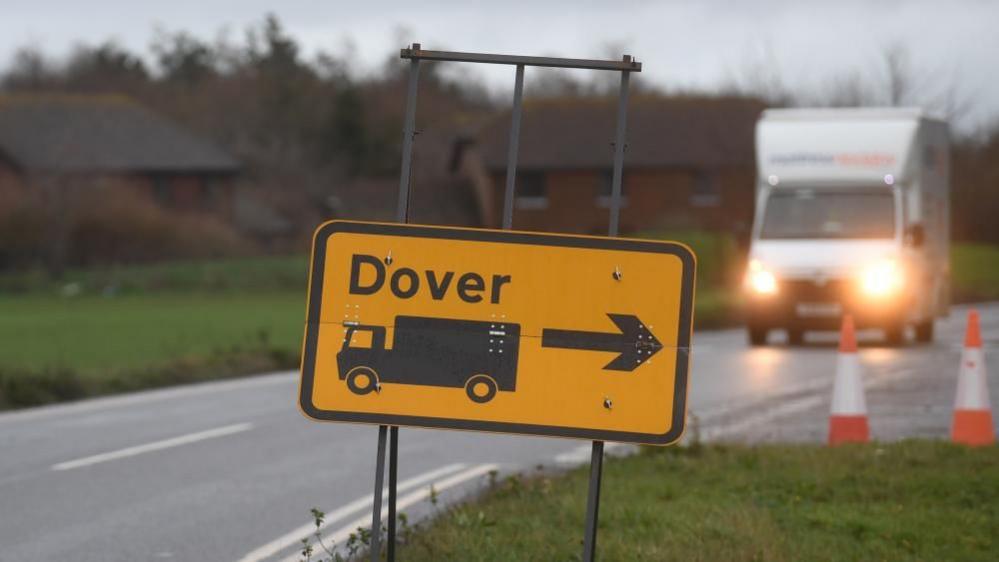 A sign for Dover: A rectangular yellow road sign sitting on a grey stand. The word "Dover" is written on the sign in black. A black arrow pointing right and a black image of a lorry can also be seen on the sign. The sign is sitting on the right-hand side of a black tarmac road. A white van with its lights on can be seen in the background.