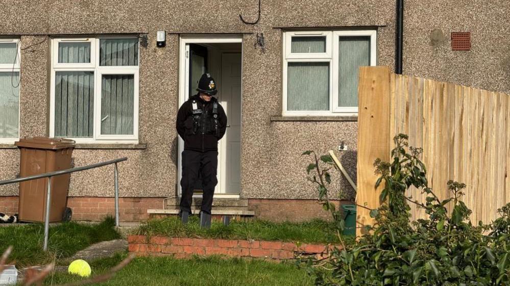 A police officer is standing in front of the door of a semi detached house. A brown wheelie bin and a yellow ball can be seen in the front garden.