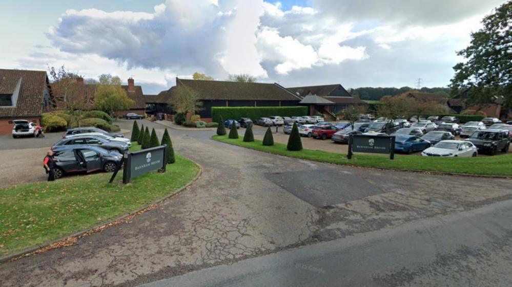 The entrance of Barnham Broom Hotel, Spa & Golf Club, showing a driveway with cars parked on both sides, bordered by grass verges. Beyond the car park is a range of buildings.