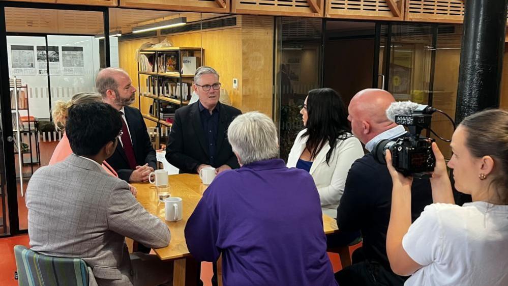Prime Minister Sir Keir Starmer, wearing a black jacket and dark blue shirt, speaks to small business leaders around a wooden table. A camerawoman can be seen filming the interaction in the foreground.