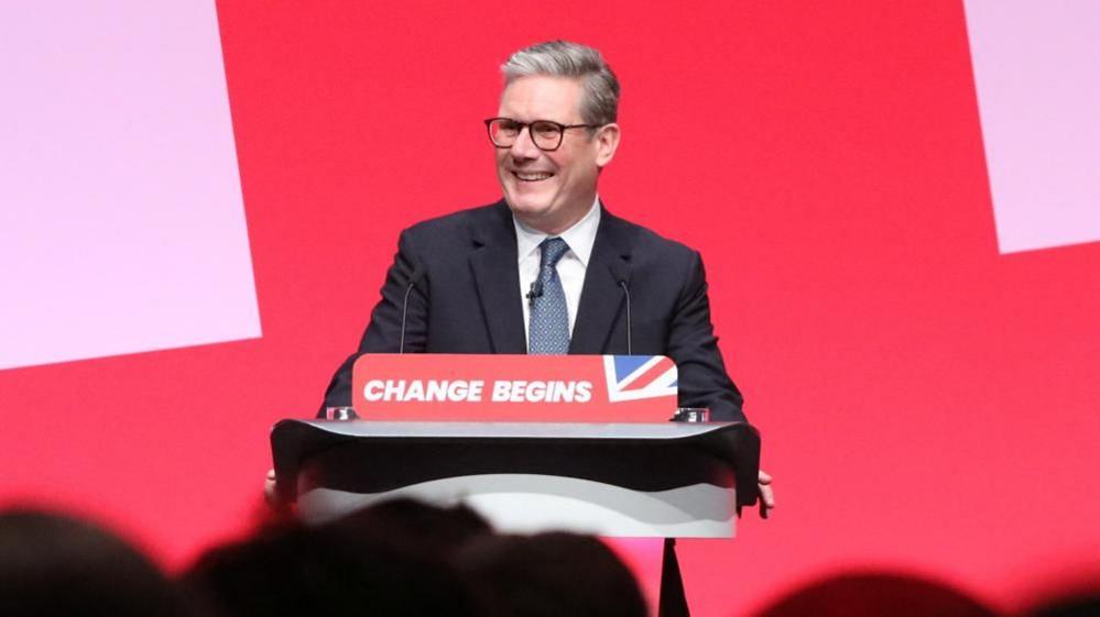 British Prime Minister Keir Starmer smiles as he delivers his keynote speech  at a lectern, emblazoned with the words "change begins".