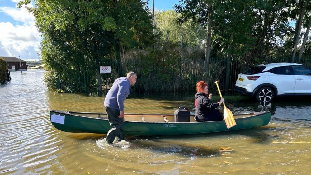 A woman with red hair is sitting in a green kayak with an oar while a man wades in the floodwater alongside.  There is a suitcase in the kayak. A white car is visible nearby.