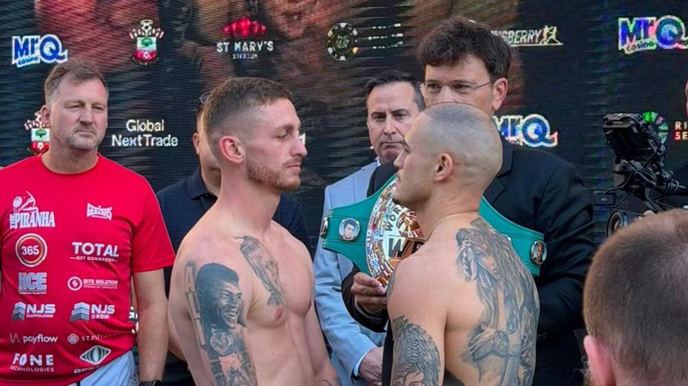 Boxers Ryan Garner and Michael Magnesi stare at one another intensely on a stage. Other people look on in the background. A championship belt is held between them by a dark-haired man in a suit. 