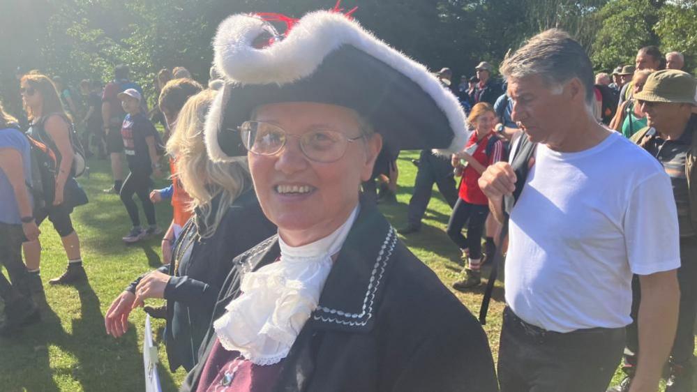 A woman with short hair and wearing glasses looks at the camera, smiling. She is part of a large group walking. She is wearing a formal hat and gown, as the town crier.