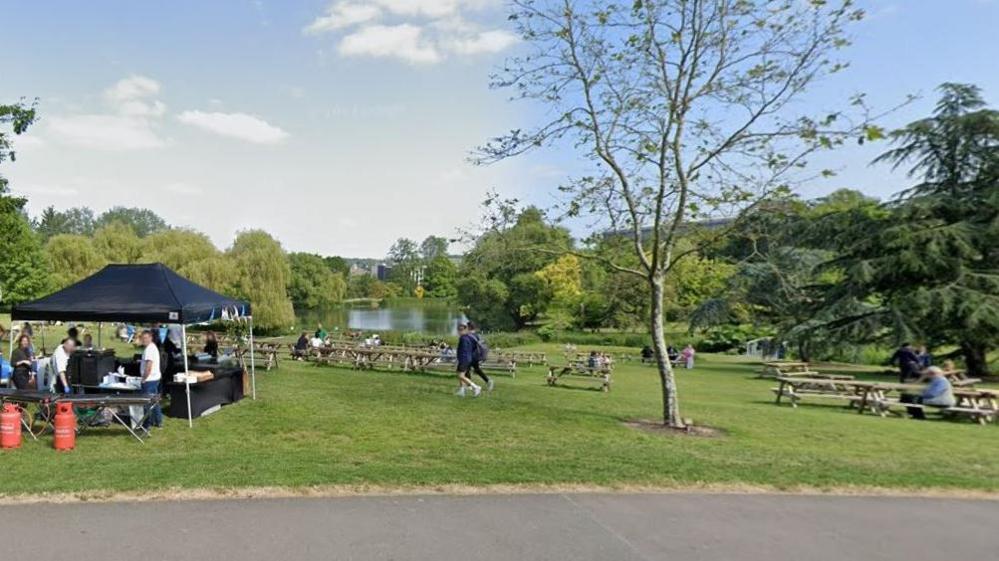 A general shot of Stoke Park in Guildford in the sunshine with people walking and sitting and a lake in the background