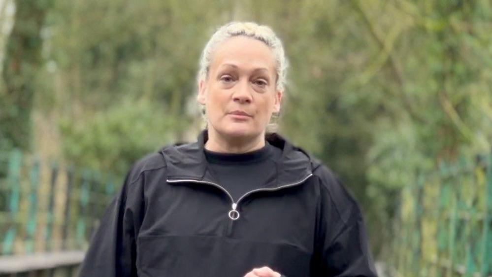 Mrs Blezard looks at the camera while standing outside. She wears a black jacket with a black top underneath. Trees can be seen behind her.