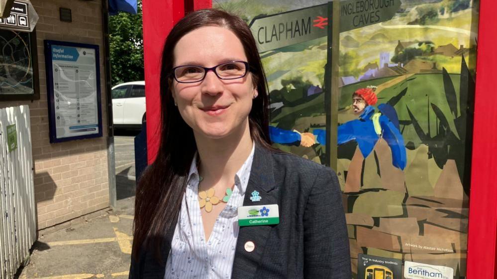 A woman with long dark hair and glasses looks at the camera. She is standing in front of a piece of artwork featuring a hiker and signs reading 'Clapham' and 'Ingleborough Caves'. She is wearing a badge reading 'Catherine' with the Bentham Line logo and a forget-me-not flower.